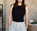 Person wearing a black sleeveless top and striped pants in an indoor setting. Round neck, pullover style, tight fitting silhouette, slim fit, ribbed knit top, sleeveless tank, rounded hem, classic, stretchy knit material.