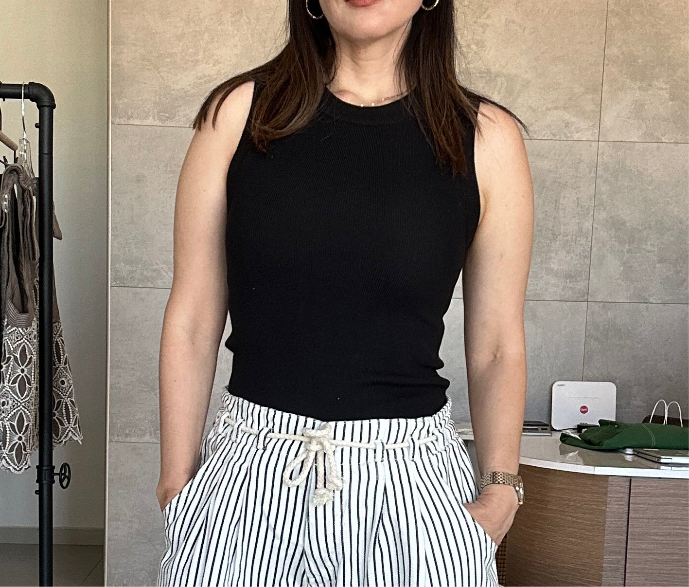 Person wearing a black sleeveless top and striped pants in an indoor setting. Round neck, pullover style, tight fitting silhouette, slim fit, ribbed knit top, sleeveless tank, rounded hem, classic, stretchy knit material.