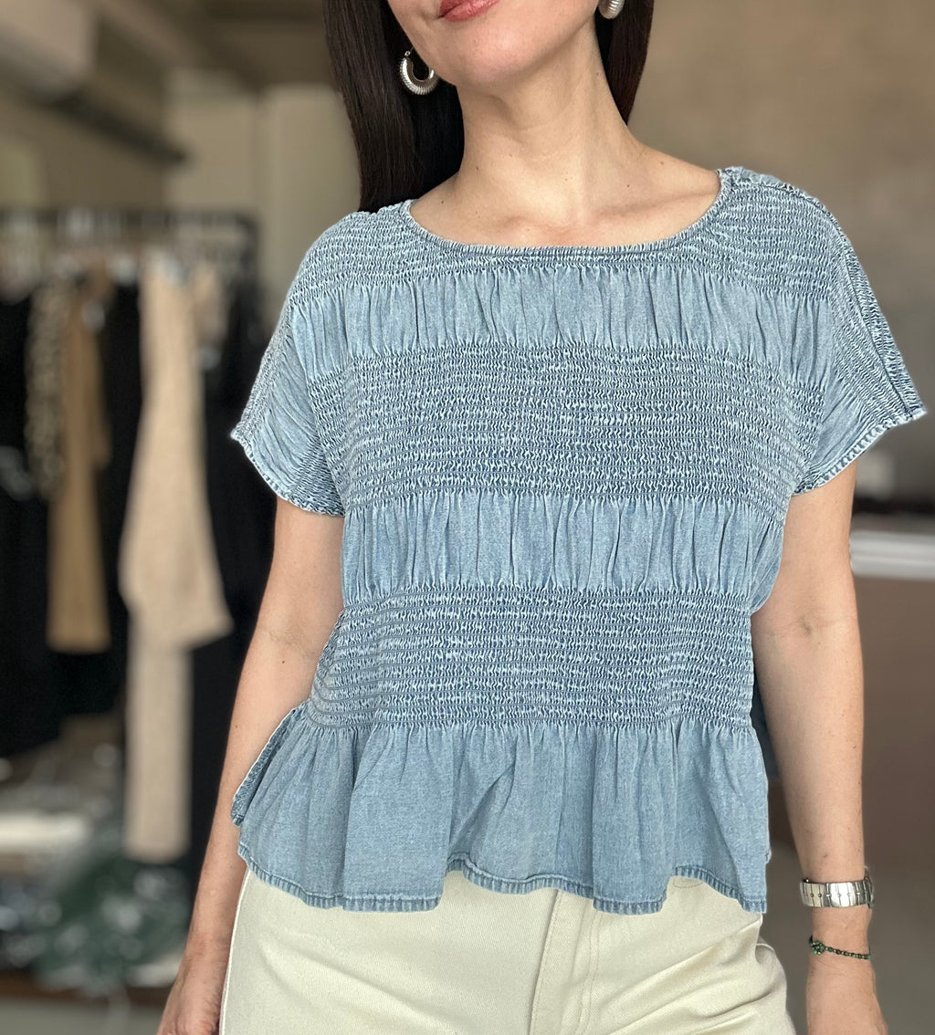 A woman wearing a light blue smocked short-sleeve top with a peplum hem, paired with cream-colored pants, in a softly blurred boutique setting.