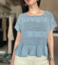 A woman wearing a light blue smocked short-sleeve top with a peplum hem, paired with cream-colored pants, in a softly blurred boutique setting.