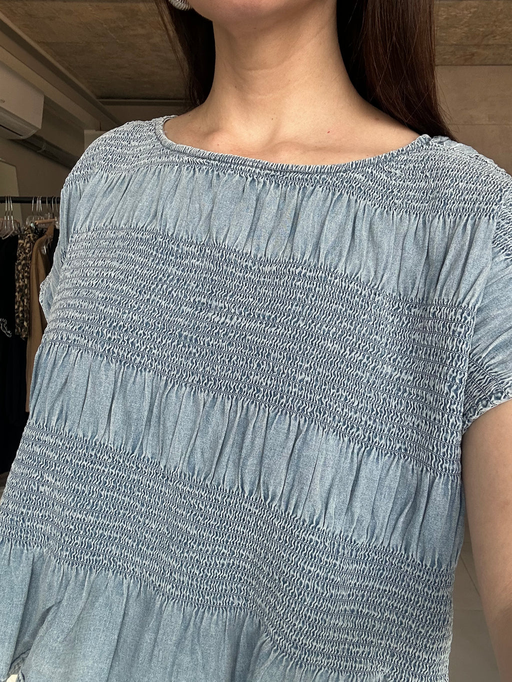 A close-up of a person wearing a light blue-gray smocked top, showing the textured fabric and shoulder area in an indoor setting.