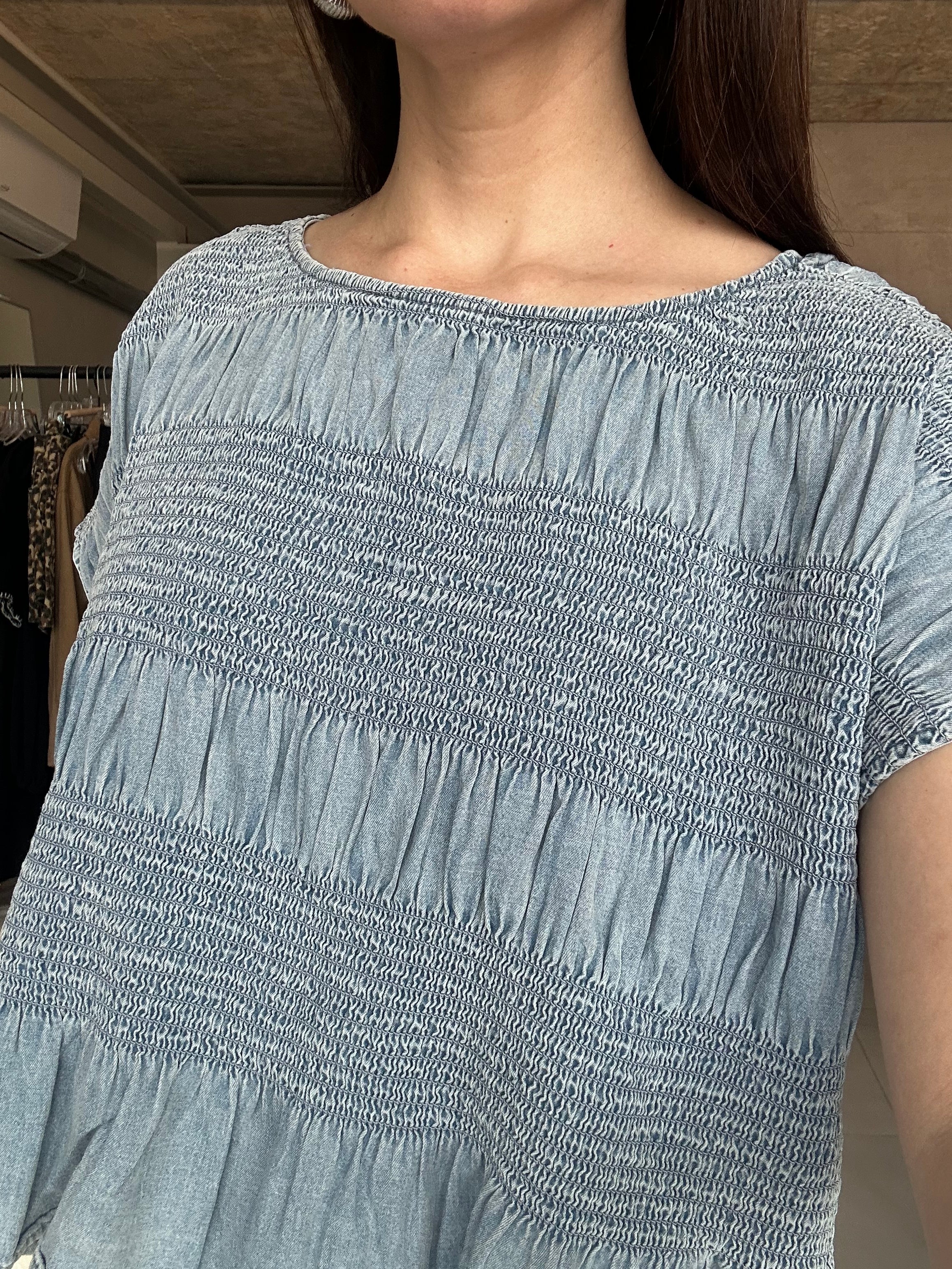 A close-up of a person wearing a light blue-gray smocked top, showing the textured fabric and shoulder area in an indoor setting.
