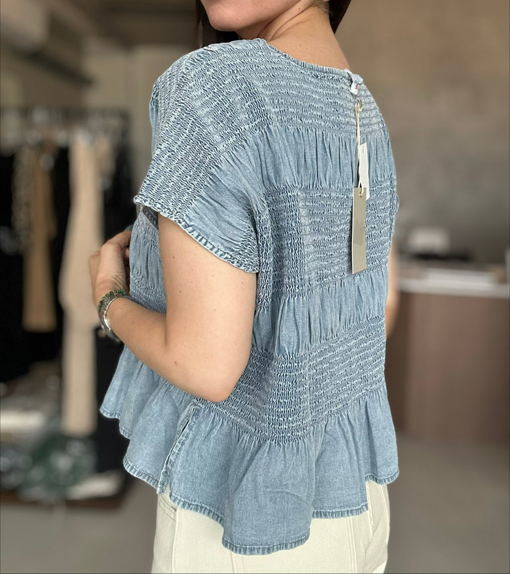 A woman wearing a light blue smocked short-sleeve blouse with a ruffled peplum hem, shown from the back in a boutique setting.
